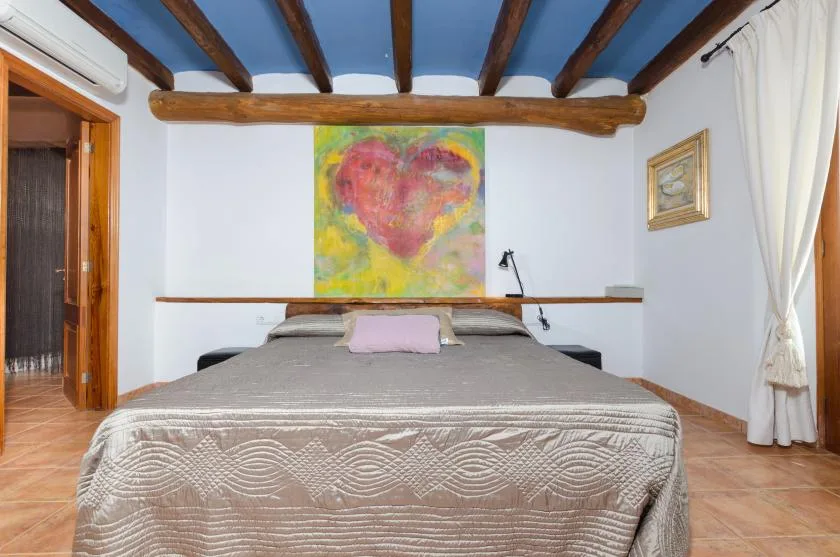 A bedroom with a neatly made bed beneath colorful wall art and exposed ceiling beams.