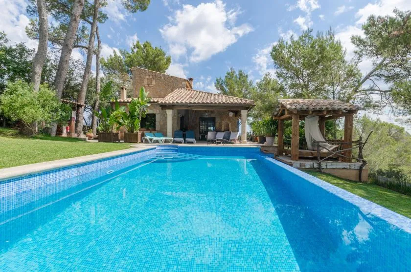 A swimming pool faces a tiled-roof house, with trees and a wooden pergola nearby.