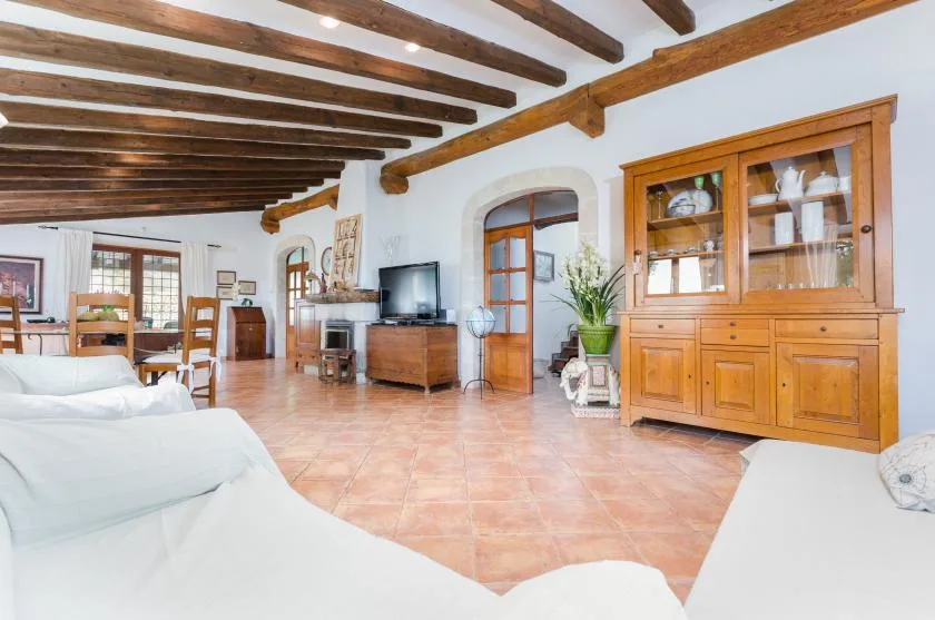 A bright living and dining area with exposed wooden ceiling beams and a glass-front cabinet.