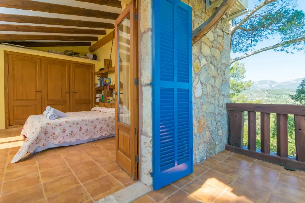 A bedroom opens onto a stone terrace with a blue shutter and wooden railing.