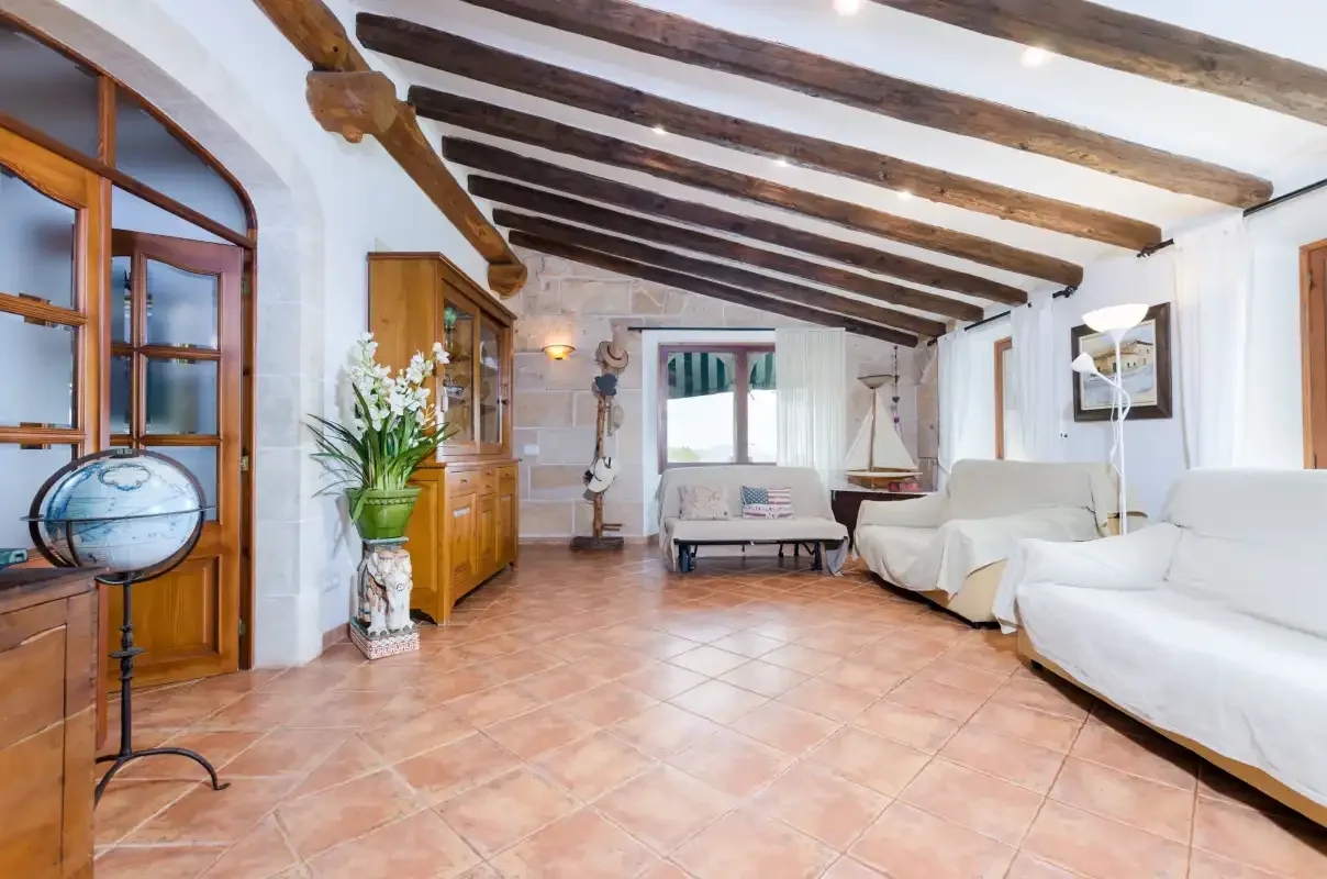 A living room with seating, exposed ceiling beams, and terracotta-colored floor tiles.