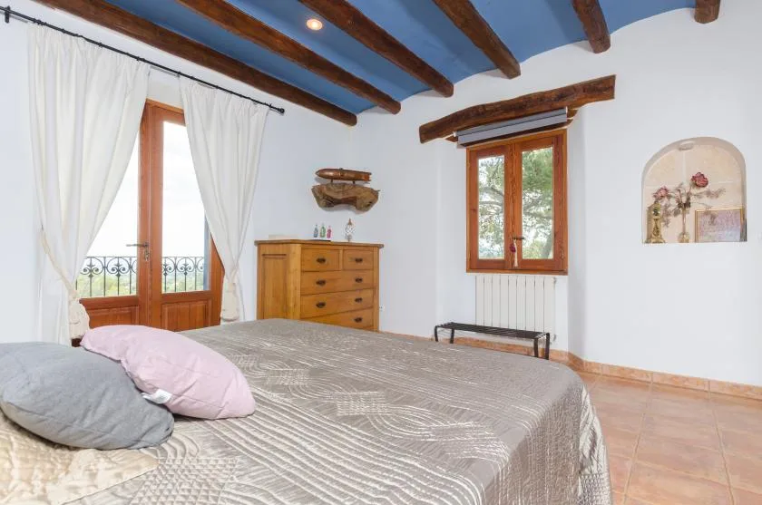 A bedroom with a bed, wooden dresser, and windows with white curtains.