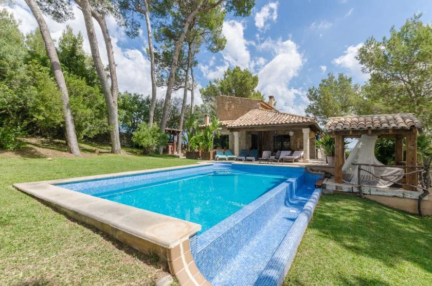 A swimming pool sits beside a tiled-roof house in a grassy, tree-lined yard.