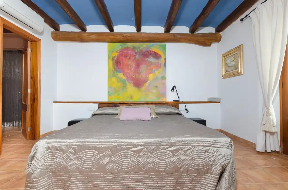 A bedroom with a bed beneath exposed wooden ceiling beams and colorful wall art.