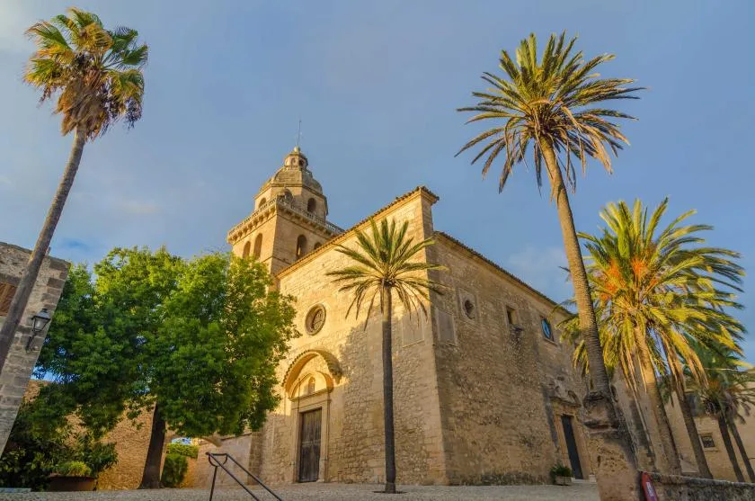A stone church with a bell tower stands among palm trees.
