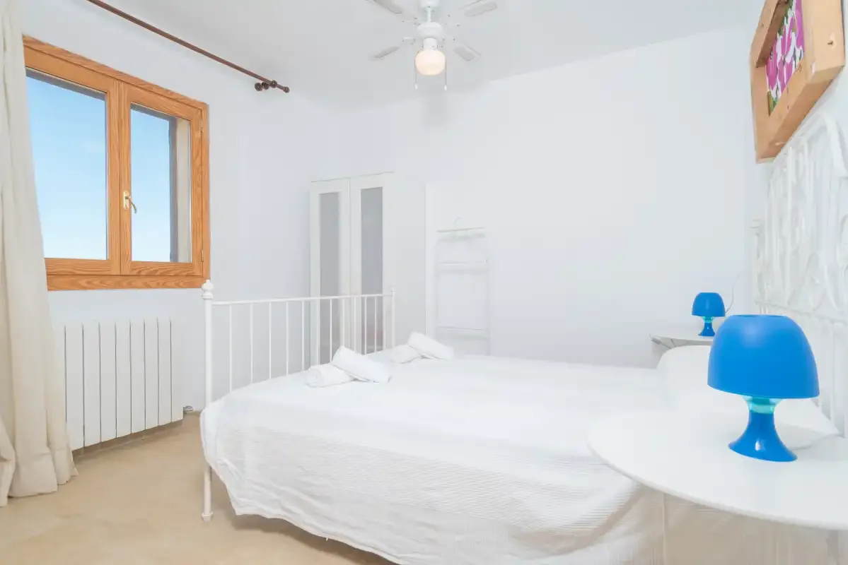 A bright bedroom with white bedding, a wooden-framed window, and blue lamps.