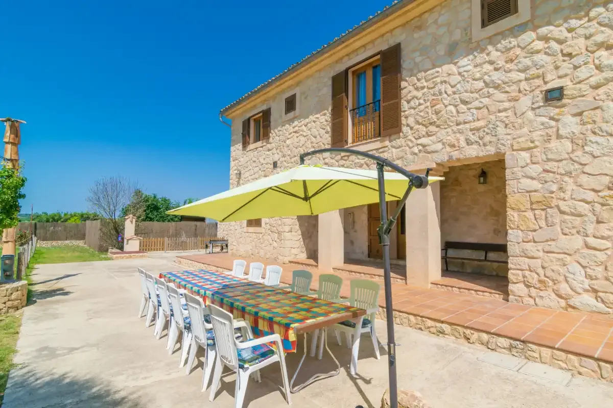 An outdoor dining area with a long table beneath a large umbrella beside a stone building.