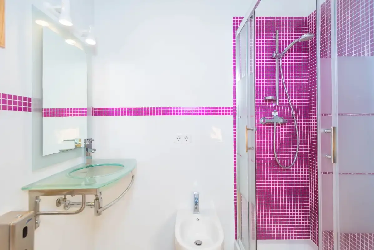 A bright bathroom with a wall-mounted sink and a pink-tiled shower.