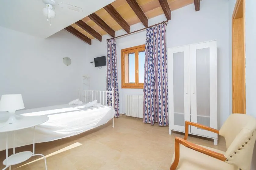 A bright bedroom with a bed, wardrobe, and curtained window beneath exposed ceiling beams.