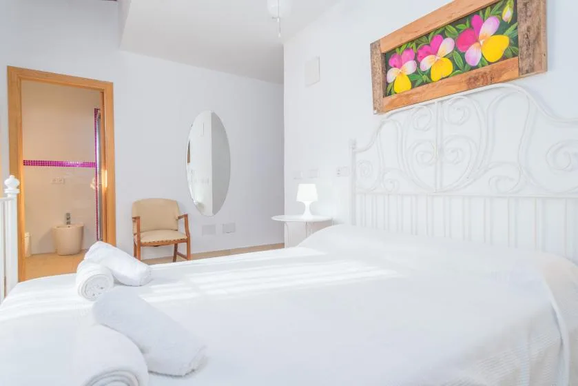 A bright bedroom with white bedding, a chair, and colorful floral artwork.