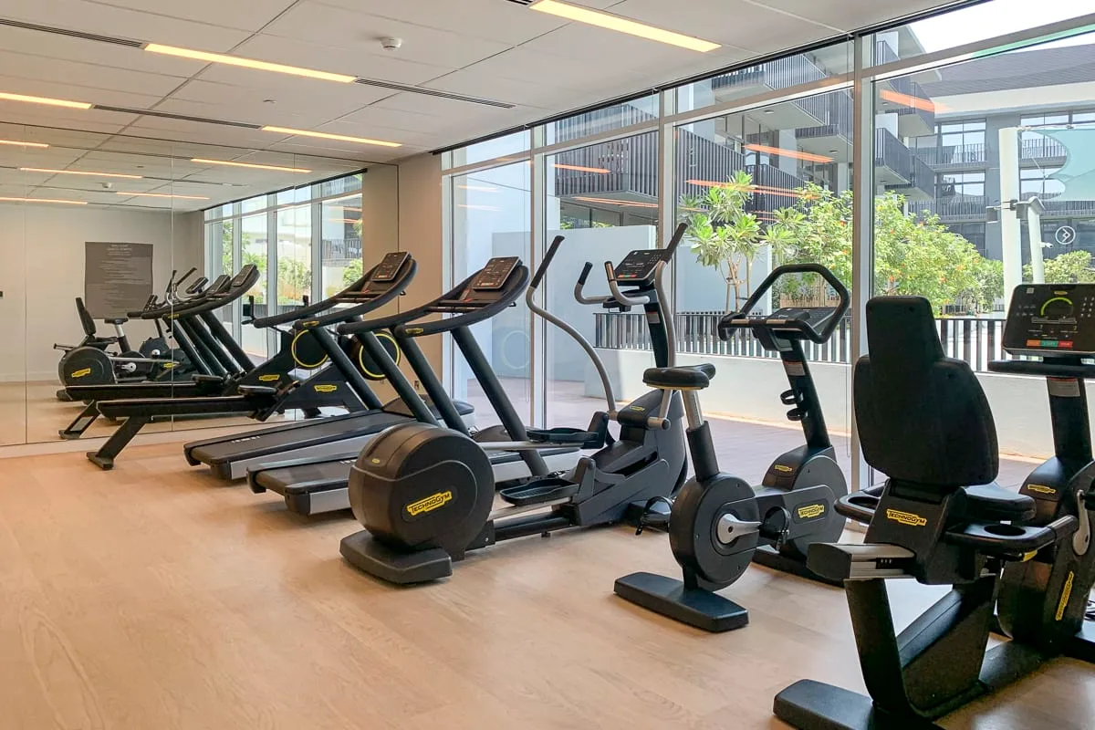 Un gimnasio con máquinas de ejercicio junto a grandes ventanas.