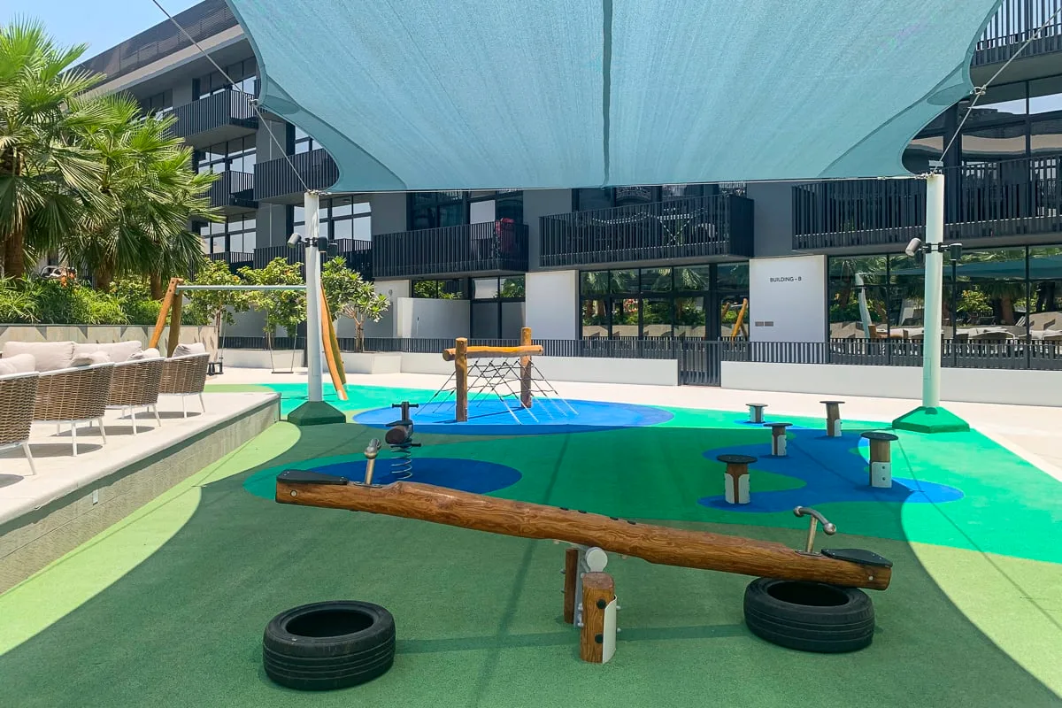Zona de juegos exterior a la sombra, con suelo de colores y una estructura de madera.