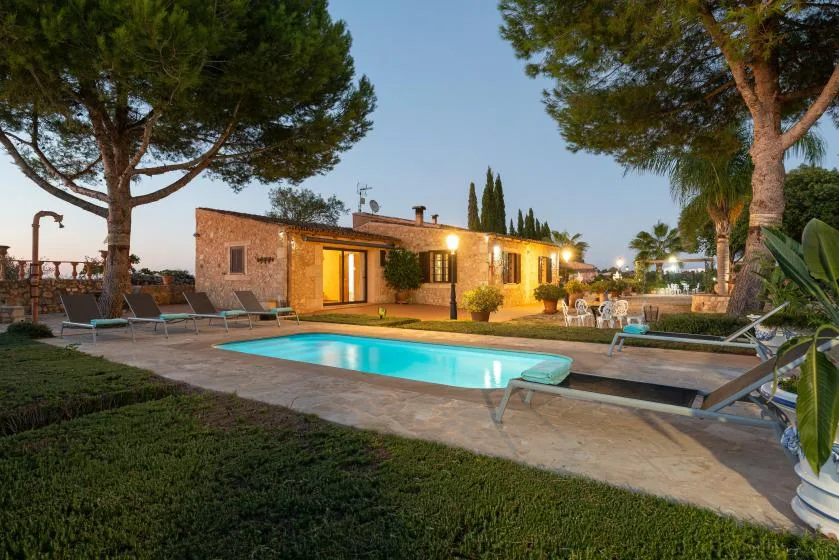 Una casa de piedra iluminada da a una piscina rodeada de árboles y asientos al aire libre.