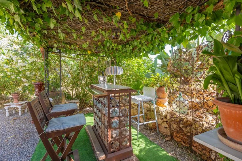 Un patio de jardín bajo una pérgola frondosa, con asientos y una mesa pequeña.