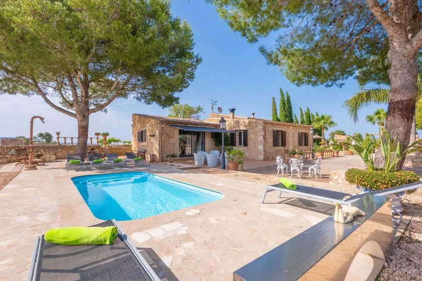 Una casa de piedra y una piscina exterior se encuentran en un patio soleado bajo los árboles.
