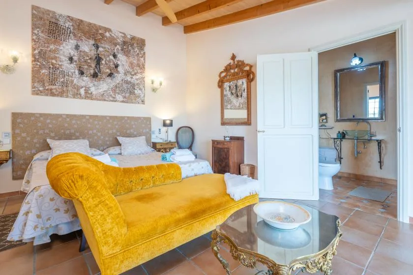 Un dormitorio con una cama, asientos amarillo mostaza y una puerta abierta al baño.