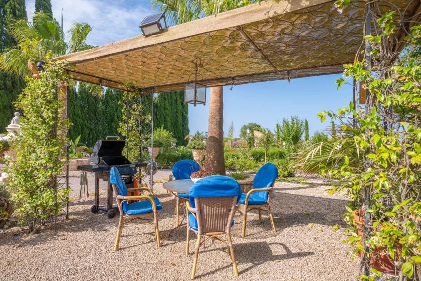 Zona de comedor exterior bajo una pérgola sombreada, con asientos azules y una parrilla.