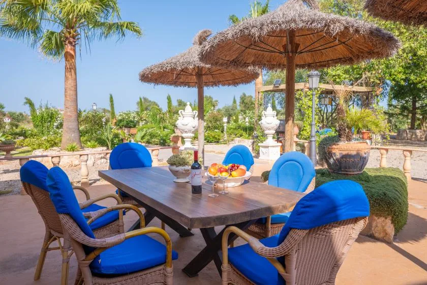 Comedor al aire libre con mesa de madera y asientos con cojines azules bajo sombrillas de paja.