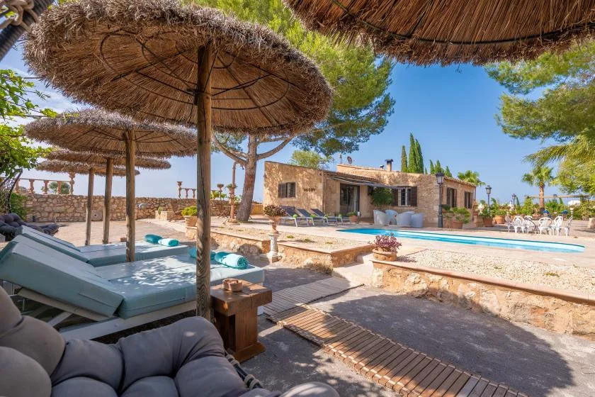 Un patio soleado junto a la piscina tiene asientos a la sombra de sombrillas de paja.