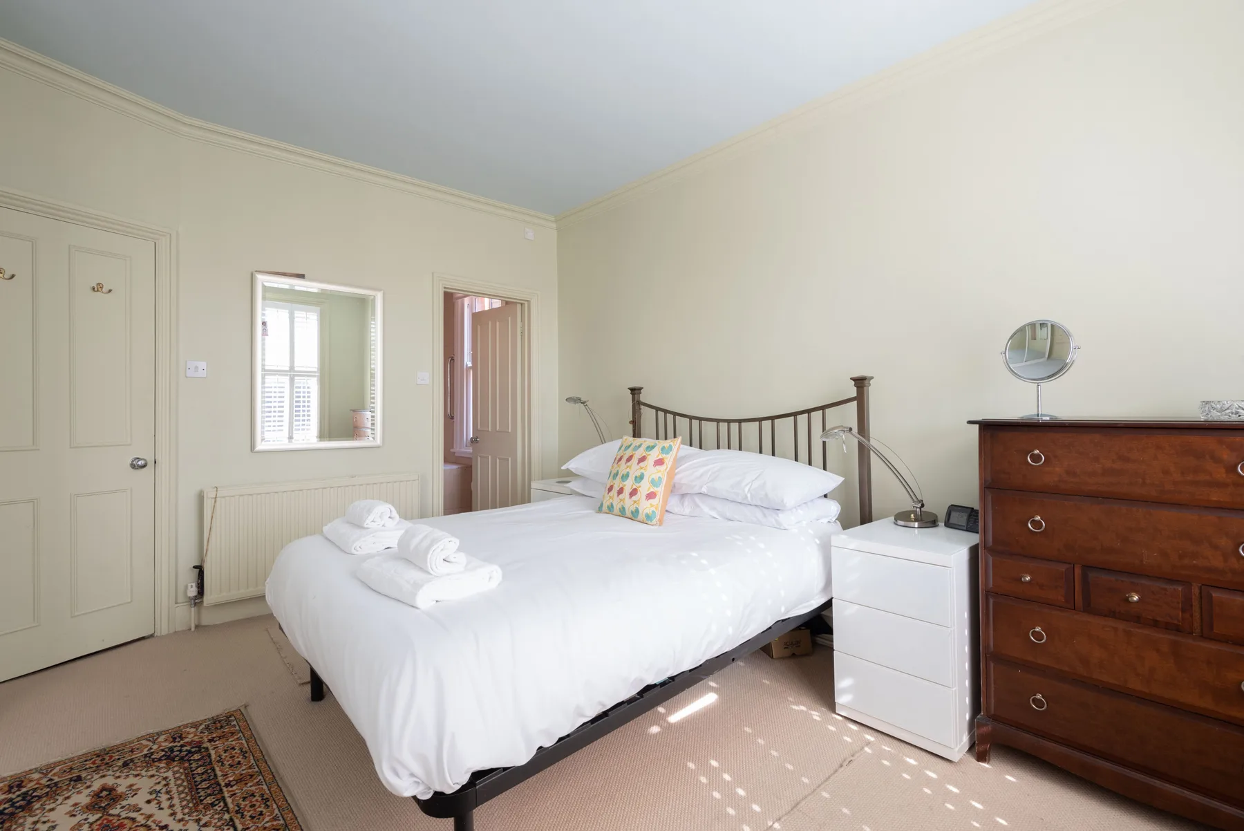 A bright bedroom with a white bed, bedside table, and wooden dresser.