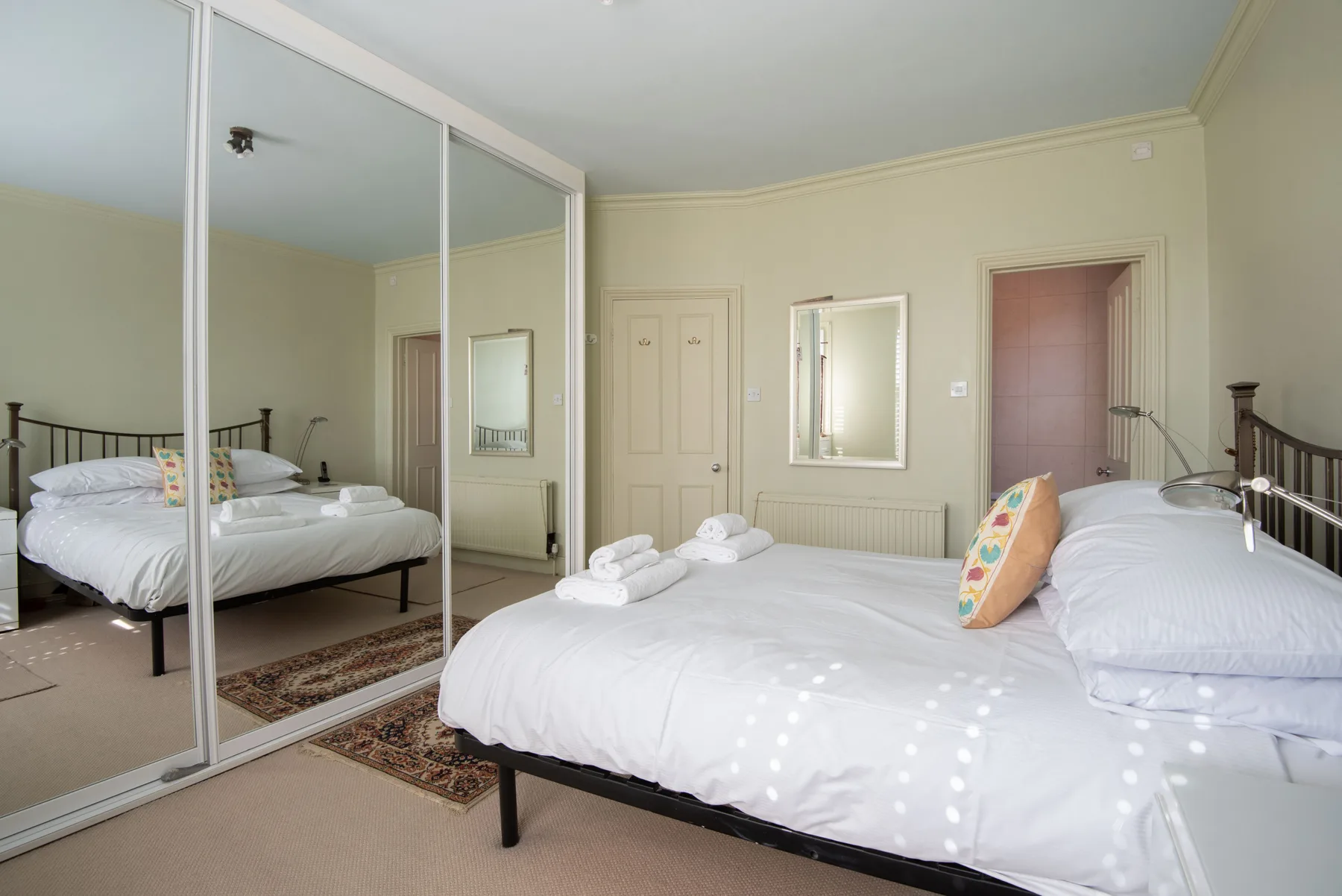 A bedroom with white bedding, mirrored closet doors, and a mirror on the wall.