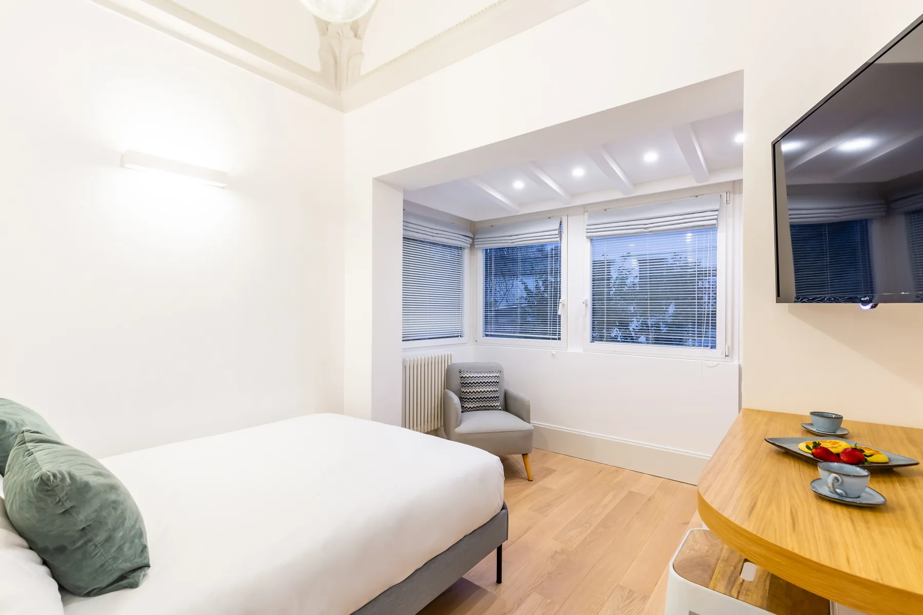 Un dormitorio luminoso con una cama, ventanas en la esquina y una mesa de madera.