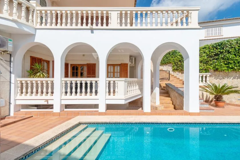 A swimming pool sits beside a white building with arched openings and a terrace.