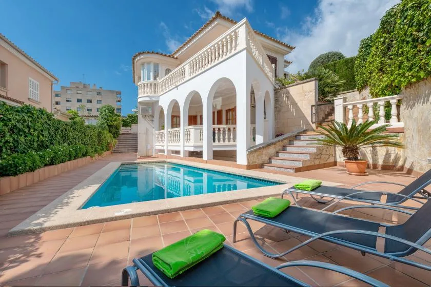 A white villa with arched verandas overlooks a swimming pool and patio seating.
