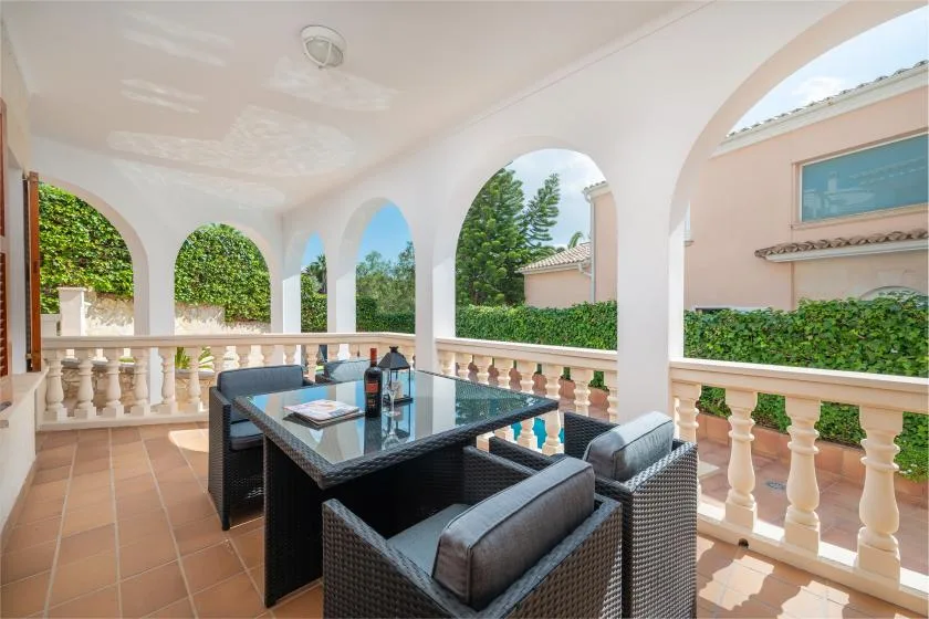 A covered terrace with a glass-topped table and woven seating overlooks a garden.