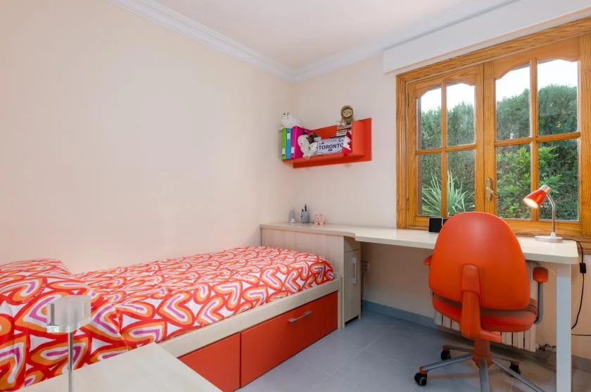 A bedroom with a bed and a desk beside a large window.
