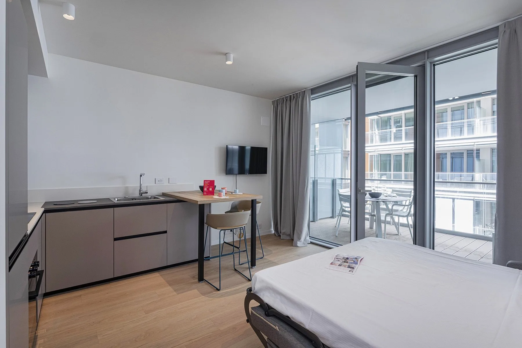 A studio room has a bed, a compact kitchen area, and glass doors opening onto a balcony.