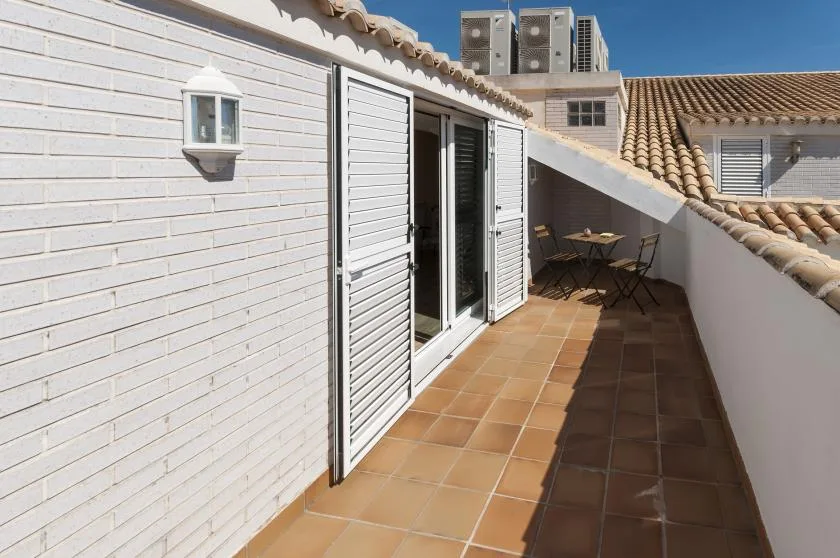 Una terraza soleada con suelo de baldosas, persianas abiertas y una pequeña mesa exterior.