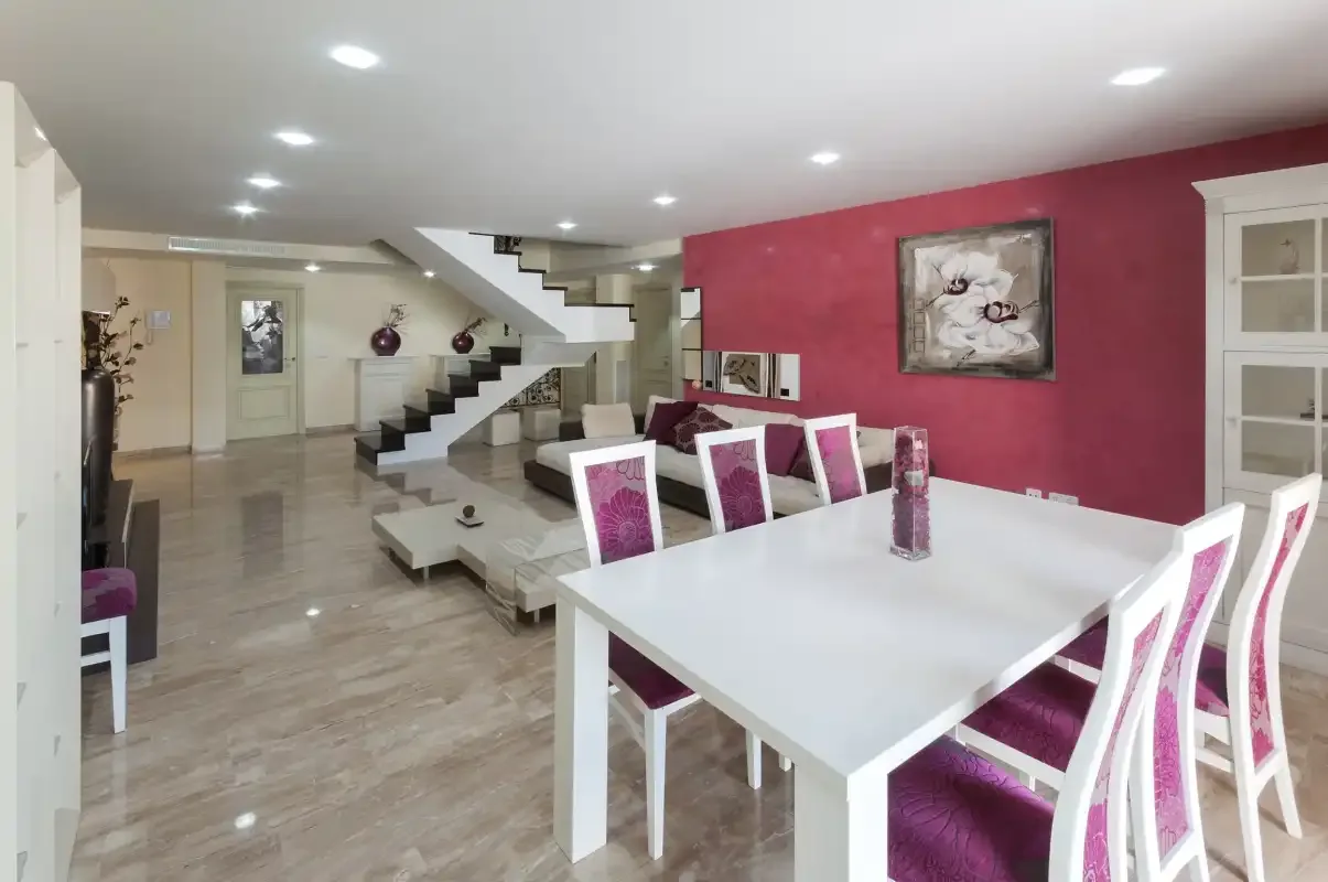 Comedor con una mesa blanca y sillas magenta junto a una escalera.