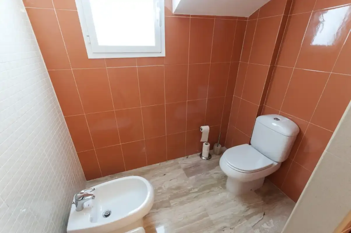 Un baño con inodoro y bidé junto a paredes revestidas de azulejos color terracota.