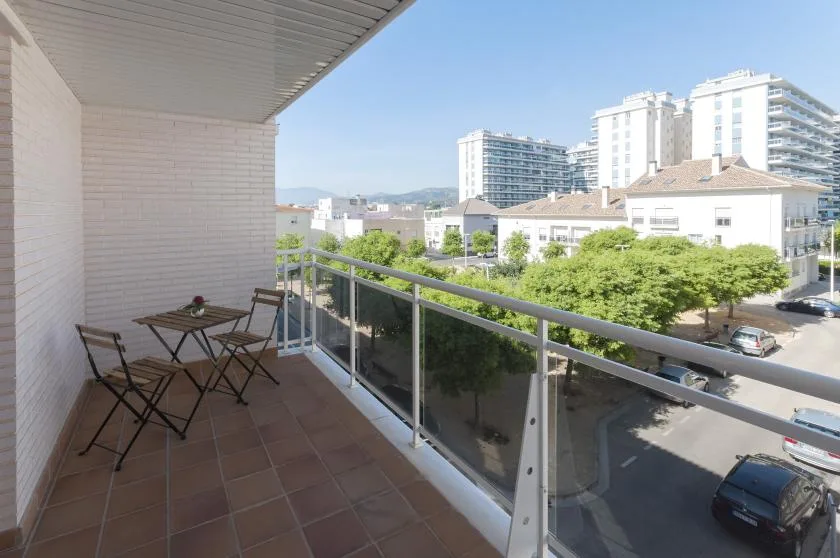 Un balcón con una mesa pequeña y asientos da a una calle arbolada y edificios cercanos.