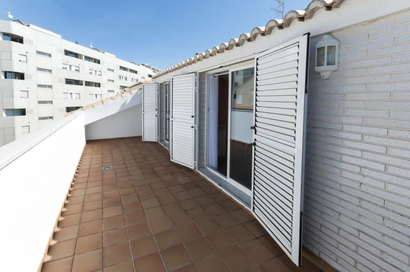 Una terraza de baldosas bordea una pared exterior blanca con contraventanas abiertas.