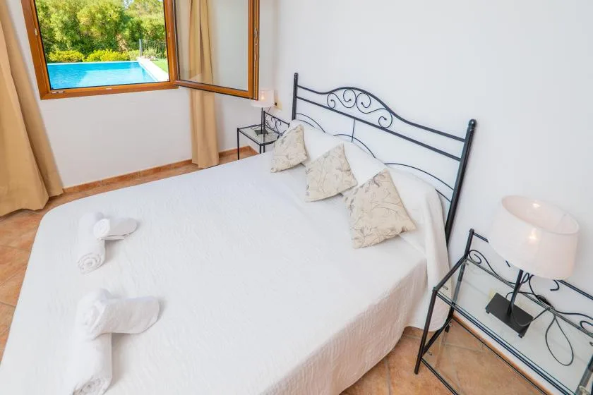 A bedroom with a neatly made bed and an open window overlooking a swimming pool.