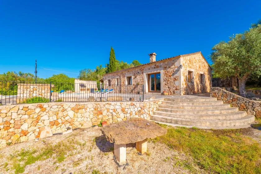 A stone house faces a broad patio with steps and surrounding trees.