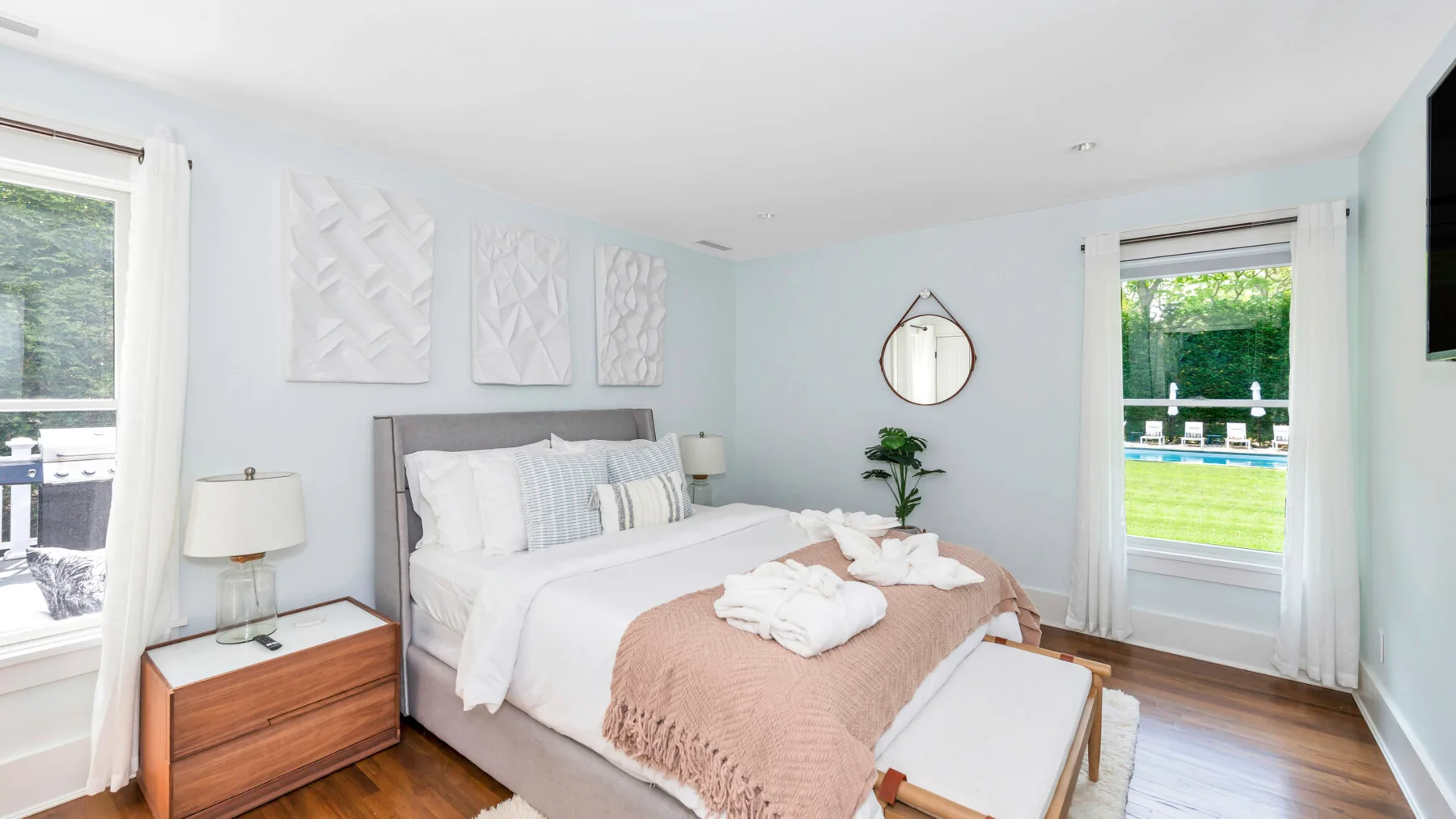 A bright bedroom with a bed, pale walls, and windows overlooking greenery.
