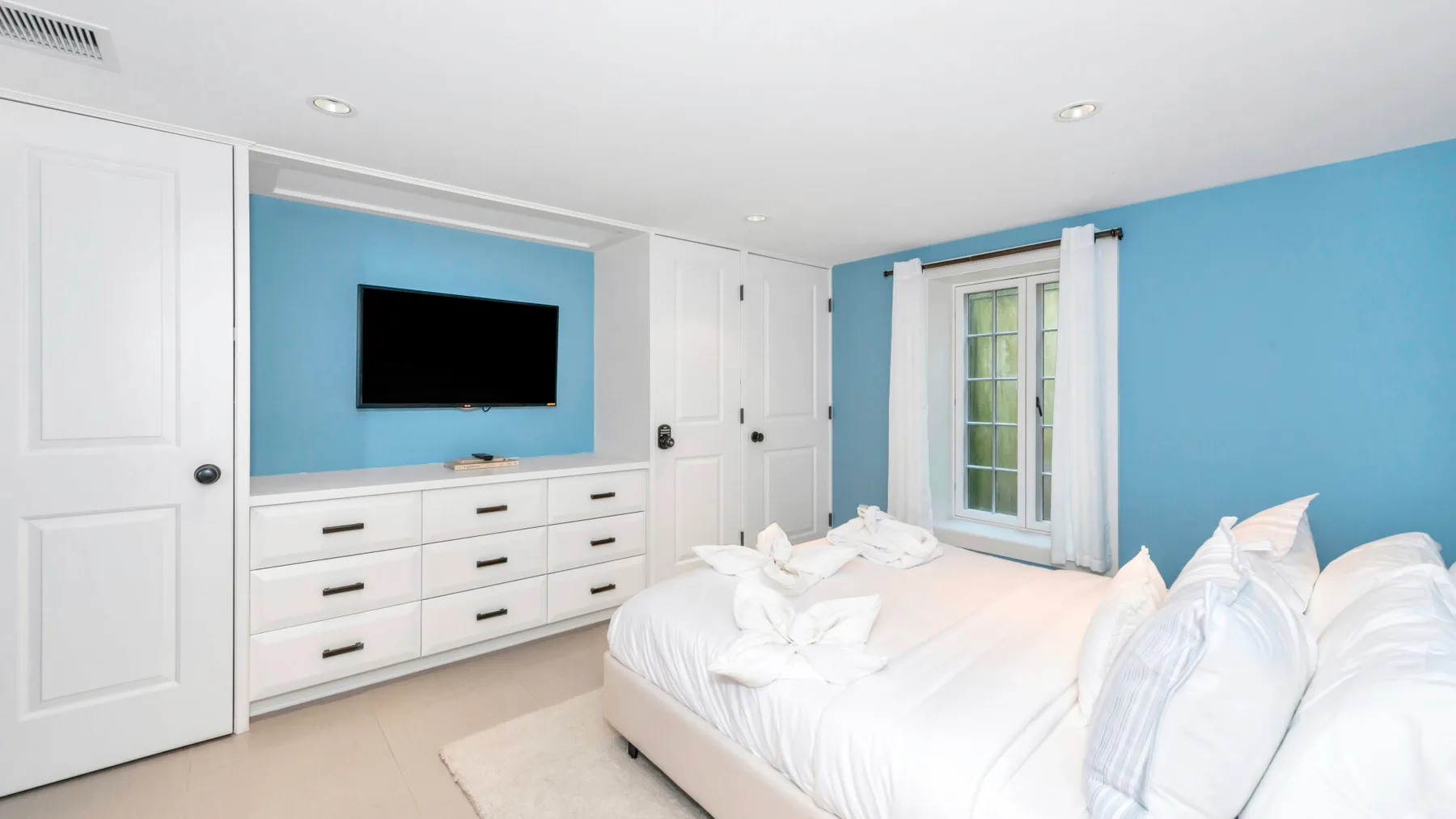 A bright bedroom with a white bed, built-in drawers, and a wall-mounted television.