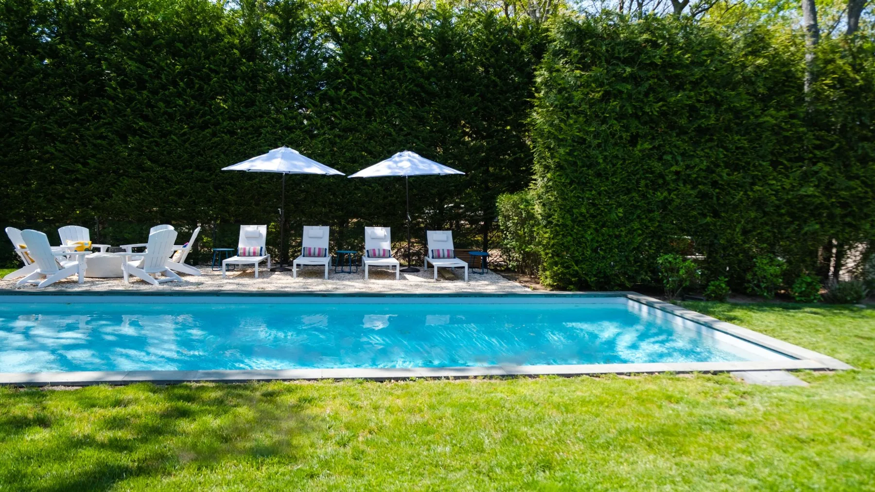 An outdoor swimming pool bordered by lawn, lounge seating, umbrellas, and tall hedges.