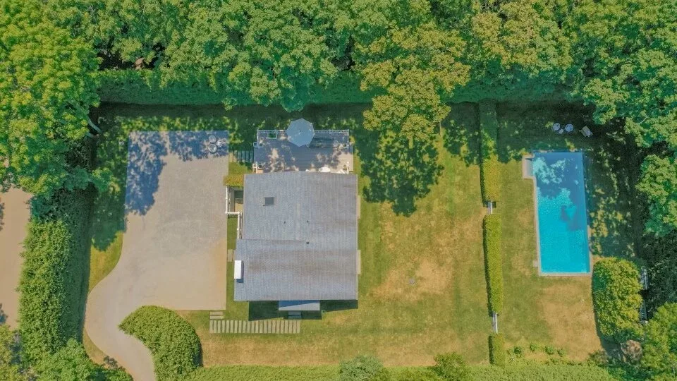 An aerial view of a house, lawn, driveway, and swimming pool among trees.