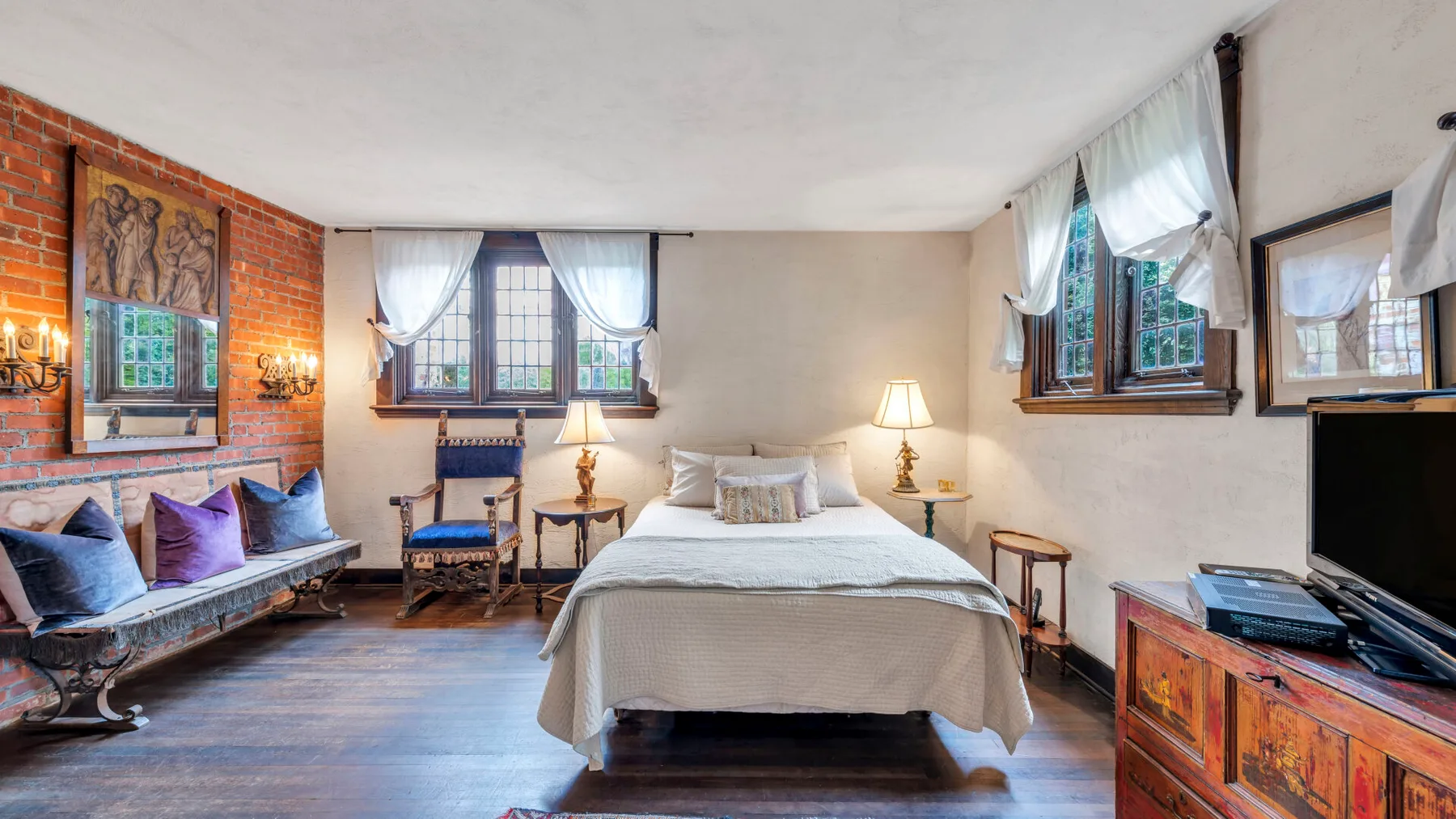 Dormitorio con una cama en el centro, ventanas con cortinas y una pared de ladrillo.