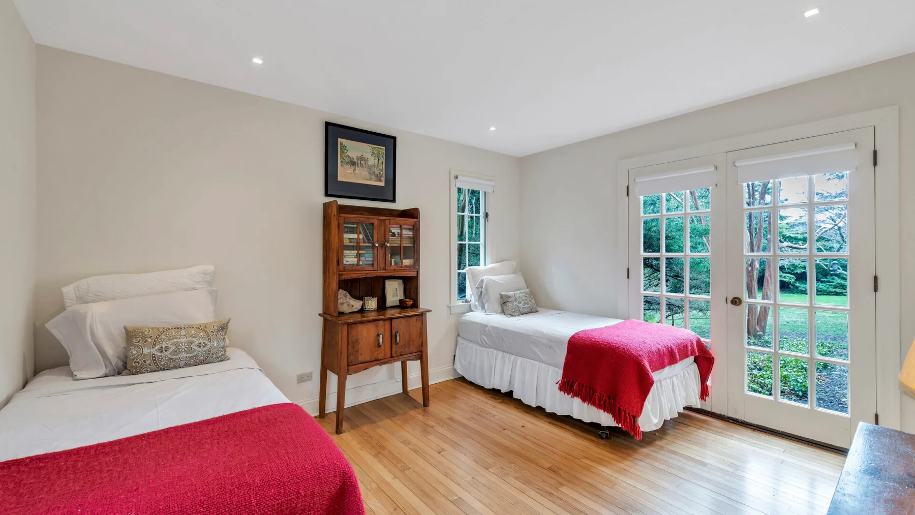 Un dormitorio luminoso con camas, un mueble de madera y puertas de vidrio hacia el jardín.