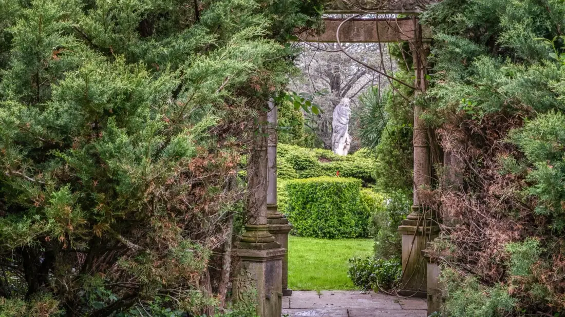 Un sendero de jardín pasa bajo un arco de piedra cubierto de hojas hacia una estatua blanca.