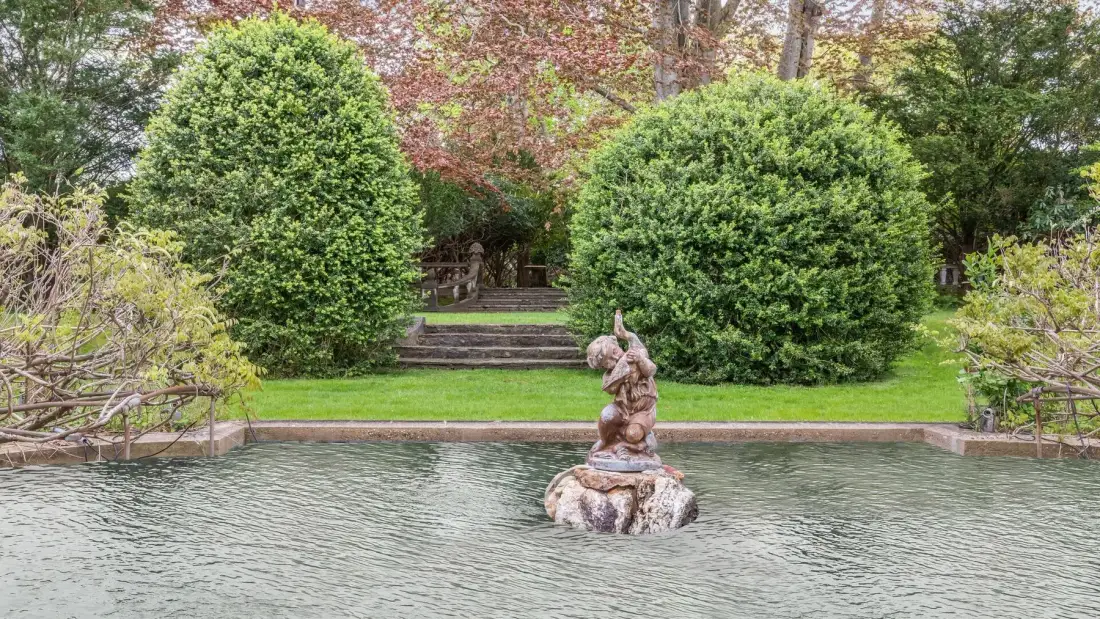 Un estanque de jardín con una estatua de piedra y vegetación al fondo.