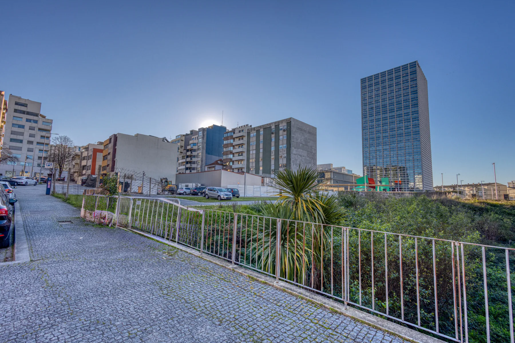 An urban streetscape with a paved sidewalk, railing, and modern buildings.