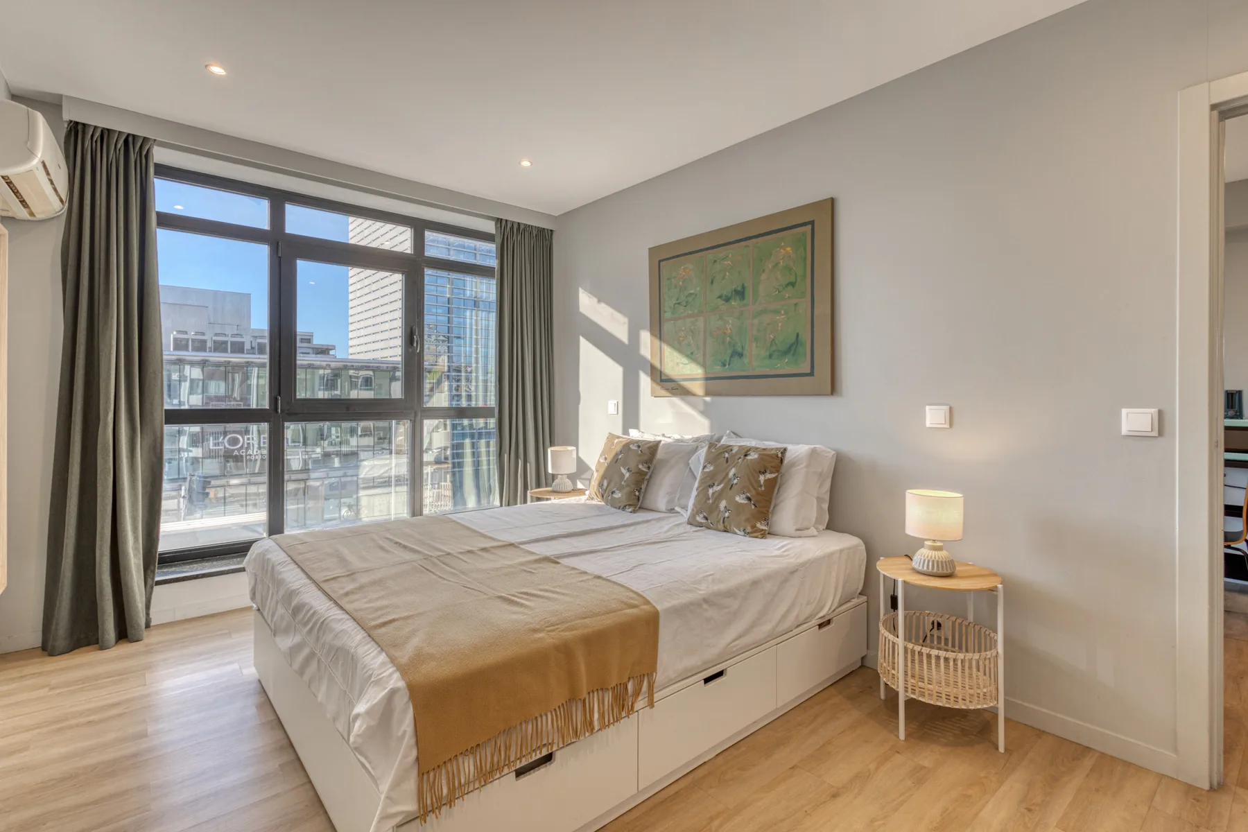 A bright bedroom with a bed beside large windows overlooking nearby buildings.