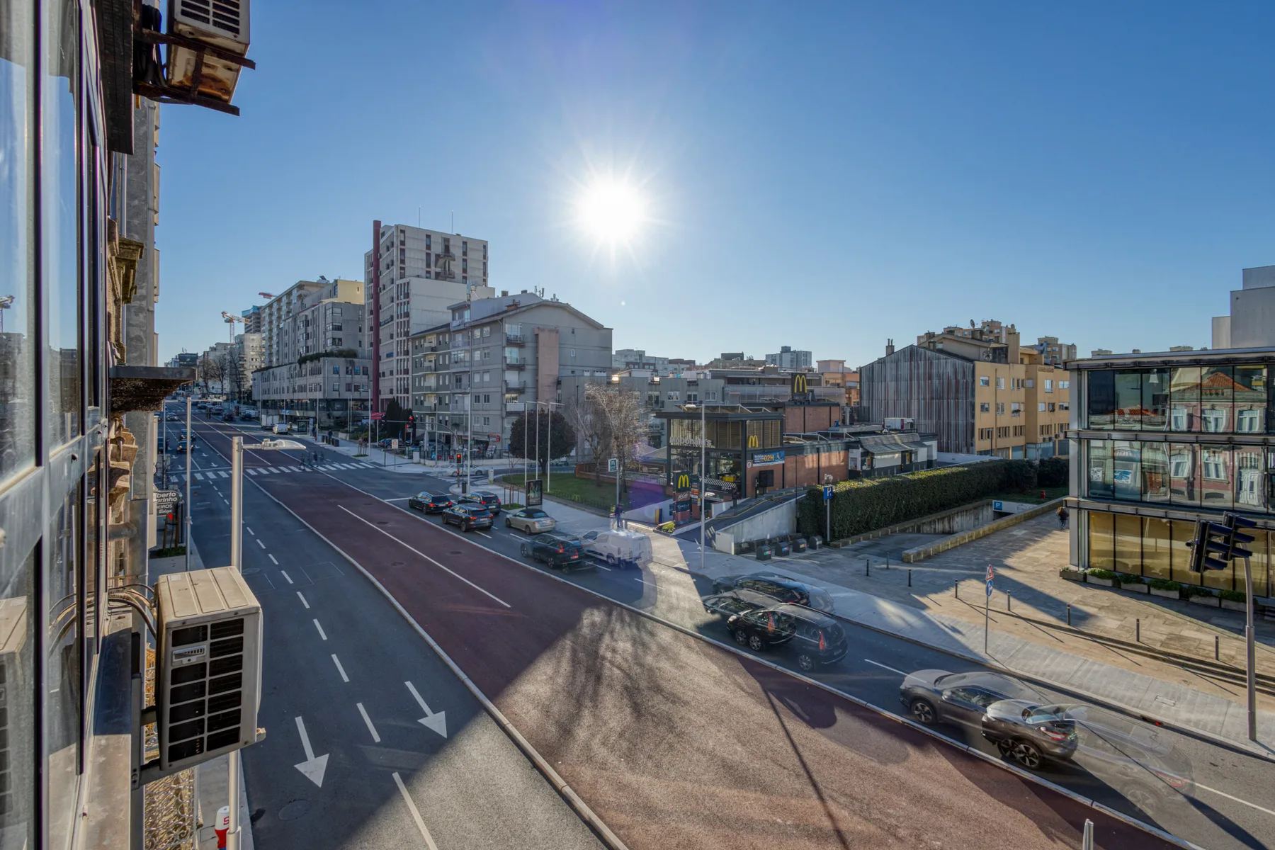 An urban streetscape with a multi-lane road, buildings, and bright sunshine.