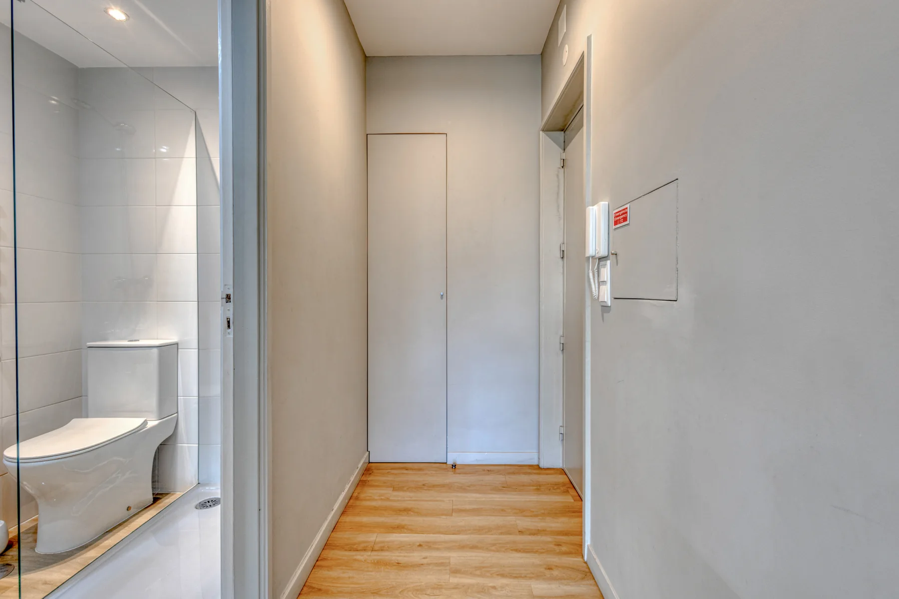 A narrow hallway with wood flooring leads to a closed door, beside a bathroom.